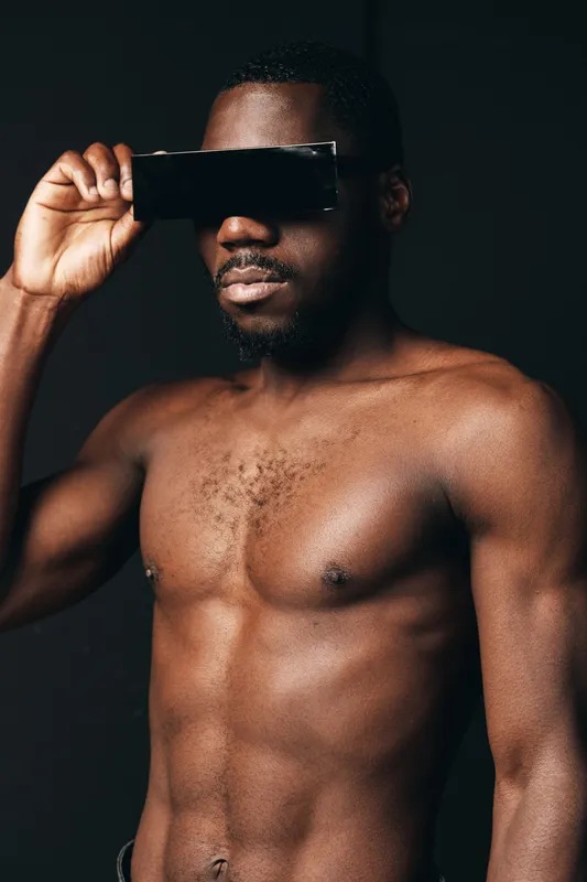African man shirtless with muscular body holding black object over eyes in creative conceptual visual style on dark background. Male model posing with serious expression and artistic lighting.