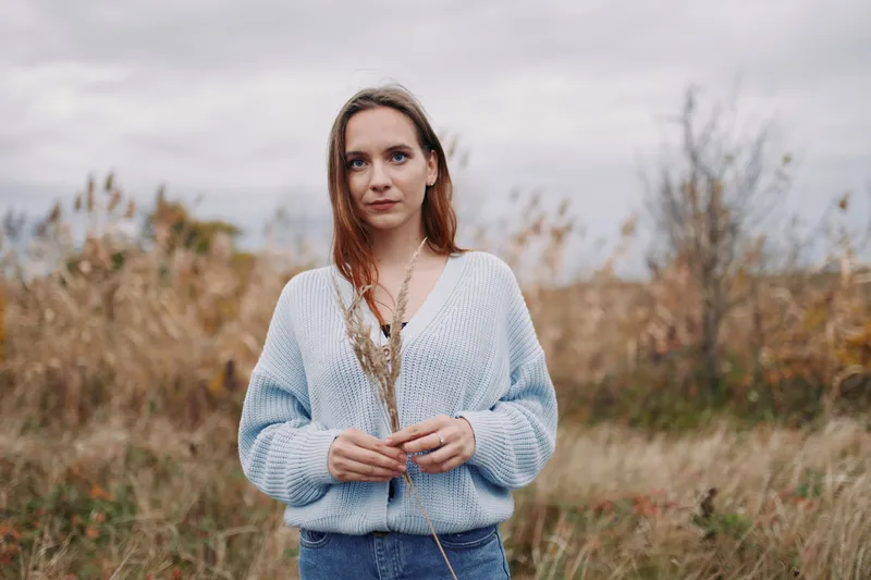 Young woman stands in a grassy field wearing a blue knit sweater, direct gaze and natural posture conveying authenticity and reliability in a candid environmental portrait under soft overcast light.