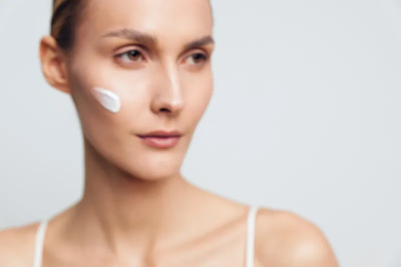 Young woman with smooth skin applying white face cream on cheek, looking away against a light background. Concept of skincare, beauty routine, health, and self-care.