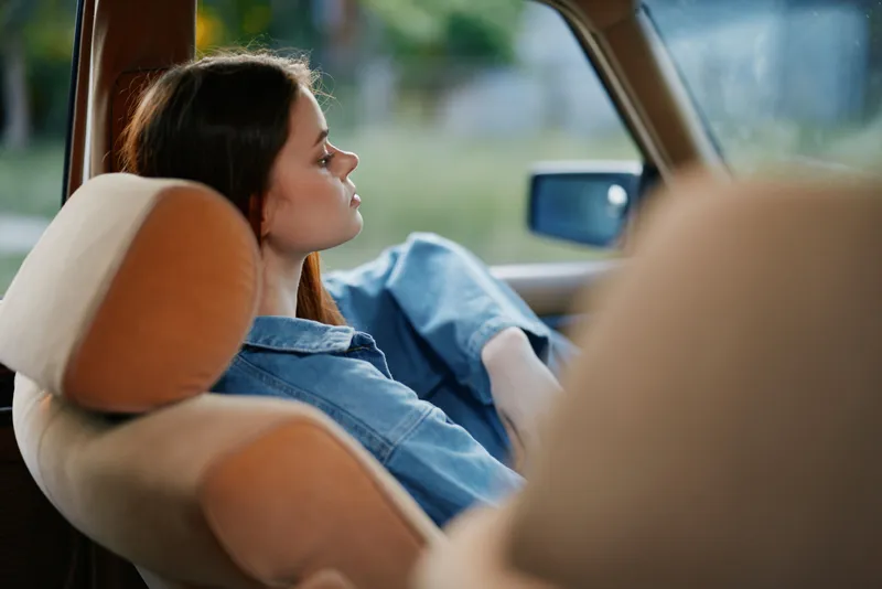 Thoughtful young woman in casual denim outfit relaxing in car, showcasing introspection and serenity on a sunny day with a serene outdoor background.