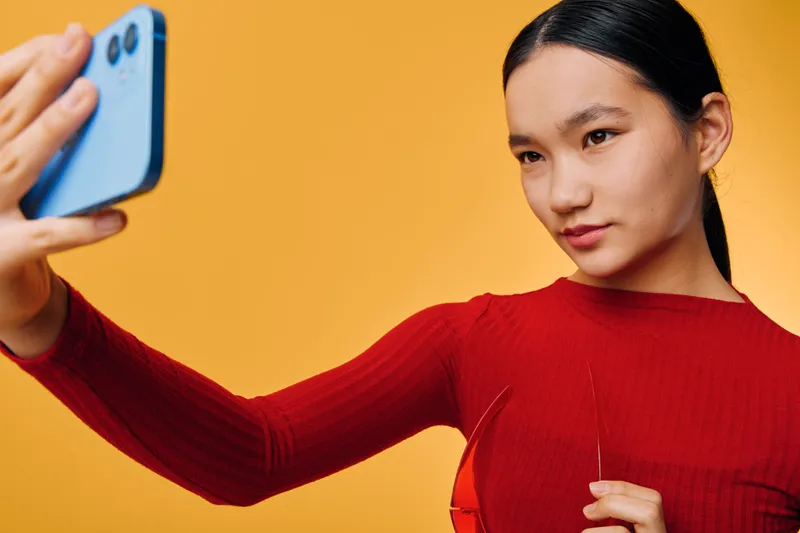 Young woman in red sweater taking selfie with smartphone, posing with focused expression against orange background for social media content and personal photography.