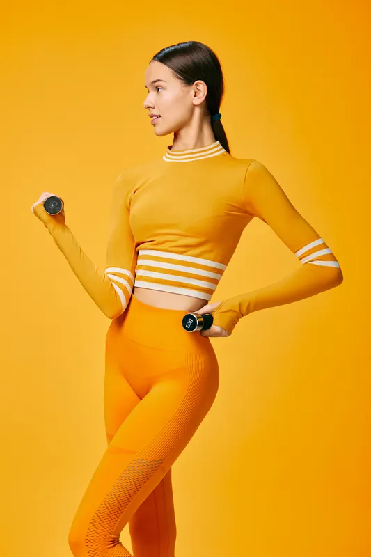 Female athlete in vibrant orange athleisure lifts dumbbells against a bright yellow backdrop, showcasing strength, focus and dynamic movement in a modern fitness moment.