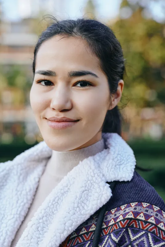 Female outdoor scene with a warm smile, wearing a patterned jacket and fuzzy fleece collar, soft natural light, casual elegant look, greenery and blurred background