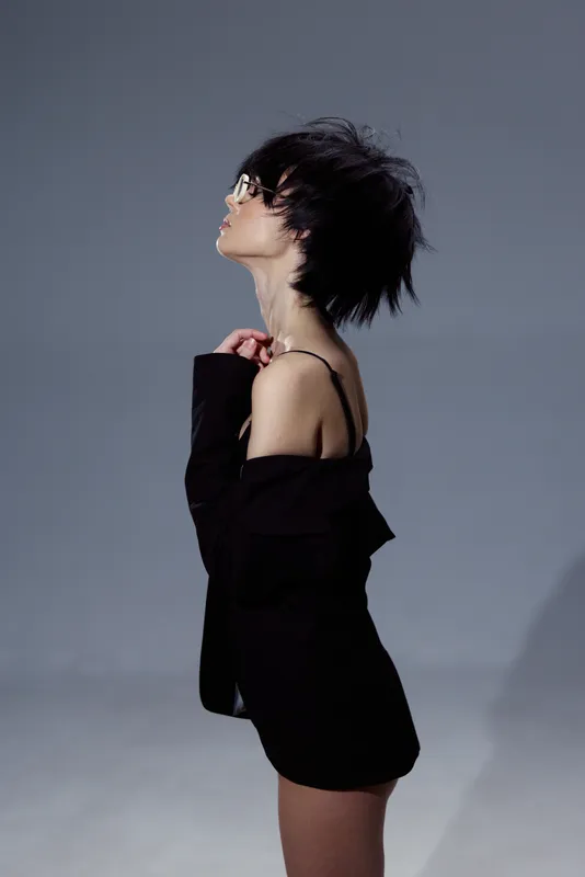 Profile of a young woman with short hair wearing sunglasses and a black off-shoulder outfit against gray background