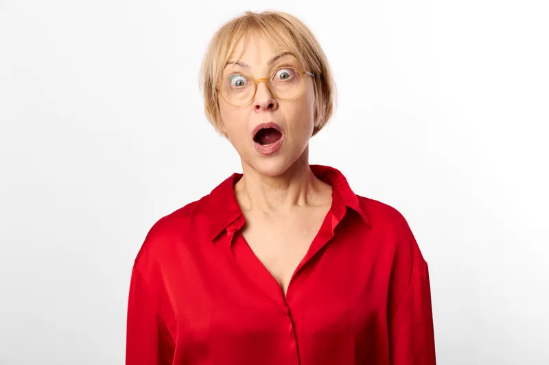 Woman with surprise and shock, shocked expression, short hair, glasses, red blouse, stares at camera, isolated, studio portrait on plain white background, high key lighting