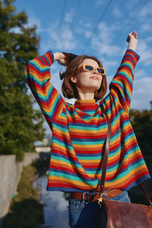 Young woman in a rainbow sweater and sunglasses, arms raised smiling on a sunny outdoor street, casual outfit with shoulder bag, joyful travel and lifestyle moment.