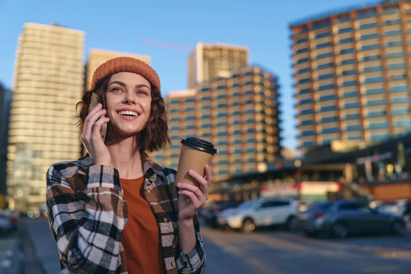 woman coffee phone city smile beanie urban portrait: candid golden hour glow scene of a smiling young woman on phone holding a coffee cup in downtown street, authenticity and mindful living.