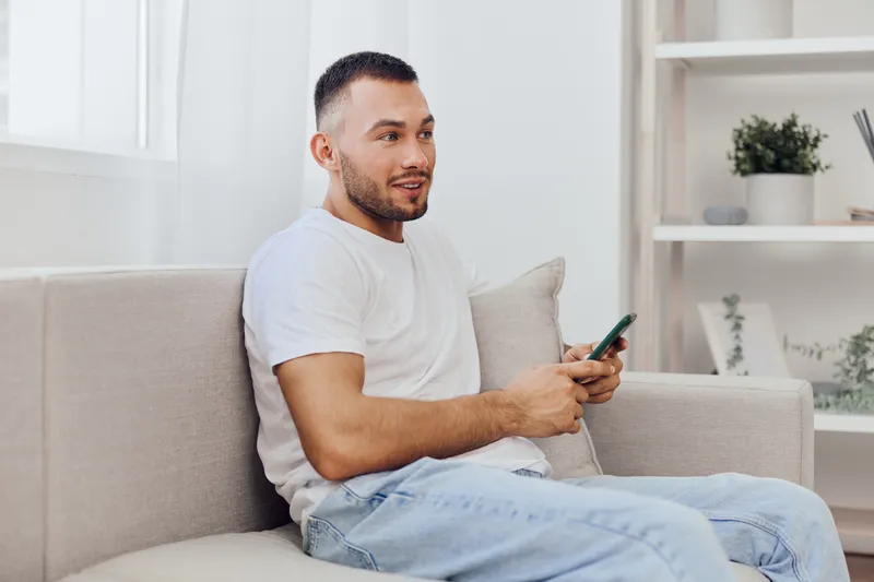 Casual man using smartphone while relaxing at home. The contemporary interior design and natural light create a cozy atmosphere that enhances modern lifestyle.