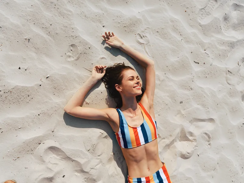 A sunlit beach scene featuring a smiling woman in a colorful striped bikini lying on warm sand, relaxed pose, casual carefree mood, summer vibe.