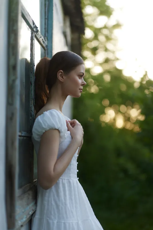 Young woman in a white dress standing by a window, showcasing serene emotion and nature in the background, ideal for seasonal wellness themes