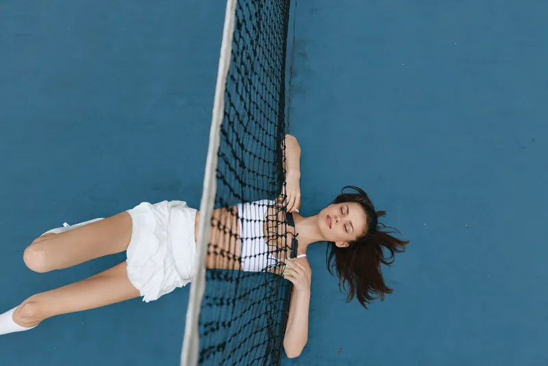 Young woman lying on a tennis court, wearing a white outfit, conveying relaxation and tranquility while enjoying a sunny day with vibrant blue background