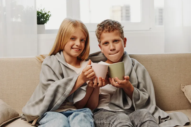 Two smiling children wrapped in gray blankets sit on a beige sofa holding mugs together in a bright room. Concept of warmth, comfort, friendship, and cozy moments.