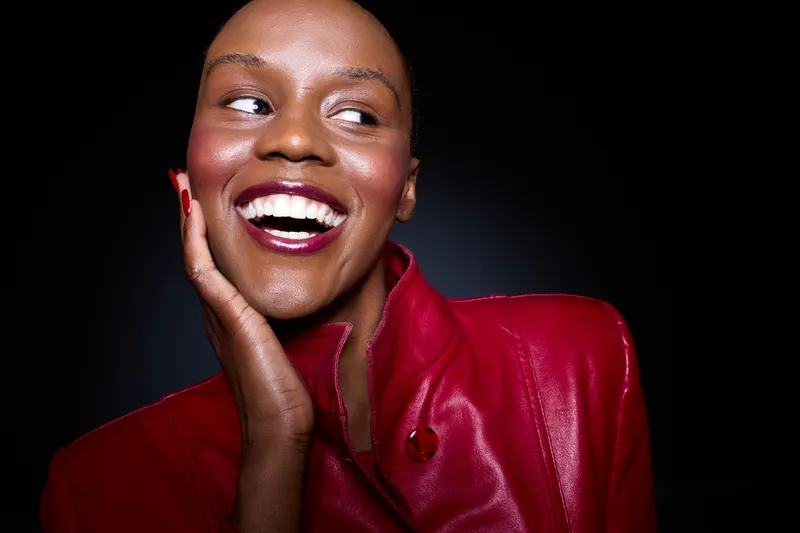 Smiling black woman with dark skin wearing red leather jacket looks to side with hand on cheek. Studio portrait with dramatic lighting creates joyful and confident mood for fashion concept.