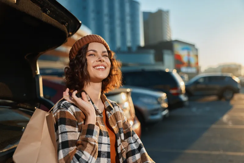Woman shopping parking sunset smile city portrait lifestyle, candid young woman with paper bags in urban parking lot at golden hour glow, authenticity and mindful living, emotional storytelling