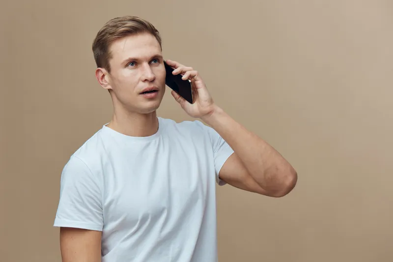 Young caucasian man talking mobile phone wear white t-shirt isolated on beige background digital nomad concept work remote technology communication portrait