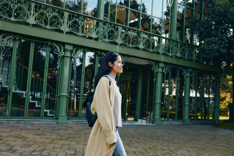 Woman walks along a paved path beside a green ornate railing and glass architecture, wearing a beige coat and backpack, urban street style and calm daytime mood.