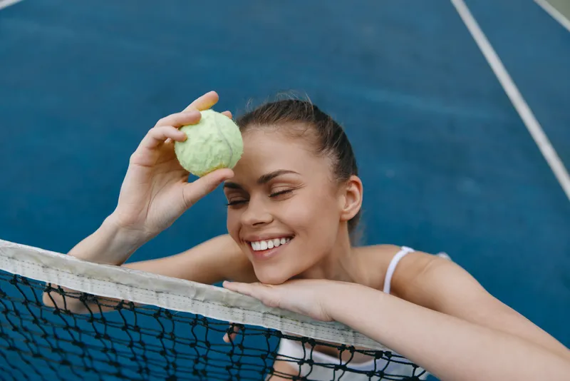 Smiling young woman holding a tennis ball by the net on a blue court, showcasing joy and enthusiasm for tennis, sporting casual attire and a healthy lifestyle