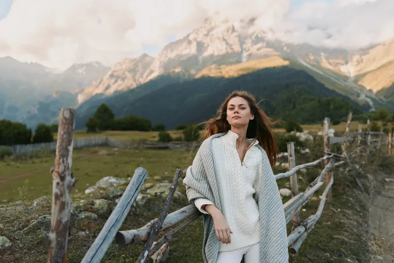 A woman in a cardigan stands beside a wooden fence in a mountainside landscape, conveying fashion driven lifestyle and relaxed portrait in outdoor setting.