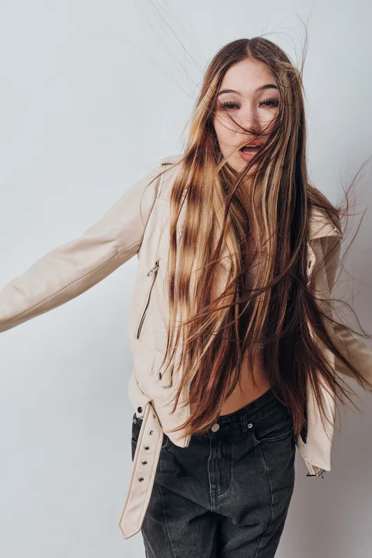 Young woman with long hair and beige jacket posing with hair blowing across face against white background. Stylish casual outfit with black pants and dynamic movement.