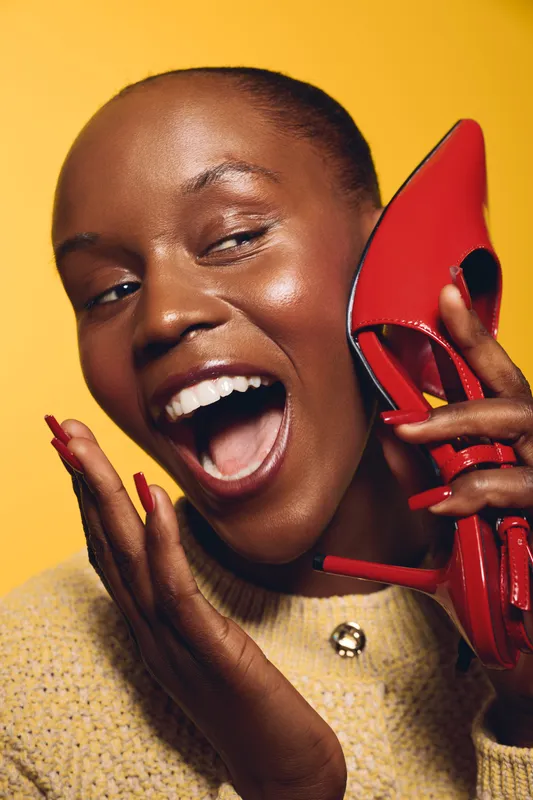 Happy African woman with dark skin holding red high heel shoe near her face, smiling widely against yellow background. Concept of fashion, beauty, joy, and lifestyle.