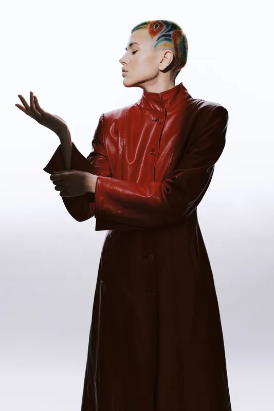 woman with colorful braided hair wearing red leather coat poses artistically on white background, showcasing unique style and confident expression in studio lighting.