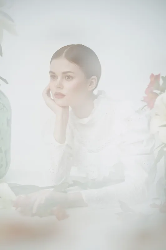 Elegant young woman in white dress, reflecting serene emotions in soft pastel background with floral elements, perfect for beauty and fashion concepts