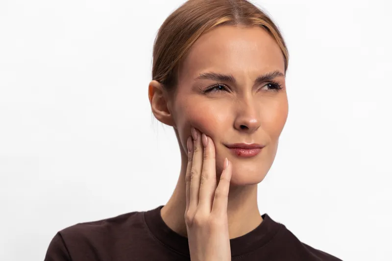 Young woman in dark shirt touches her cheek with hand, showing facial pain or discomfort. Close-up portrait on white background. Concept of toothache, dental pain, and health awareness.