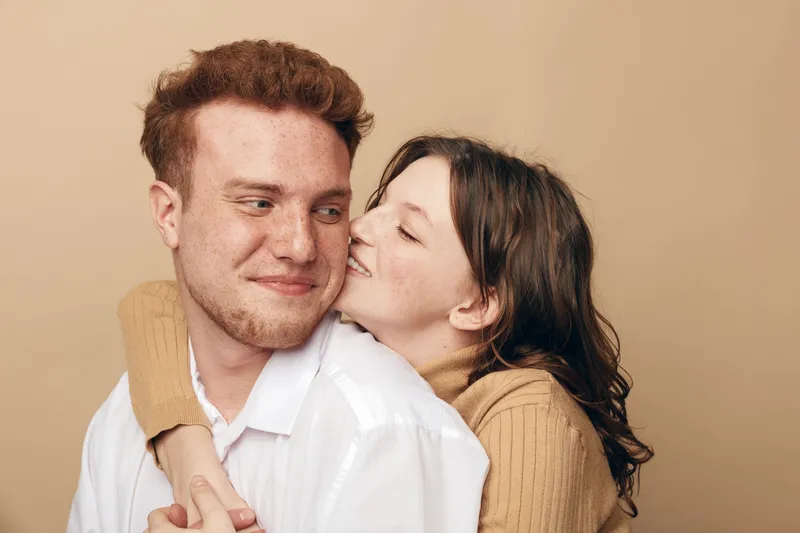 Young couple in casual clothes embraces and smiles gently against beige background. Woman hugs man from behind showing affection and warmth. Concept of love, intimacy, and relationship.