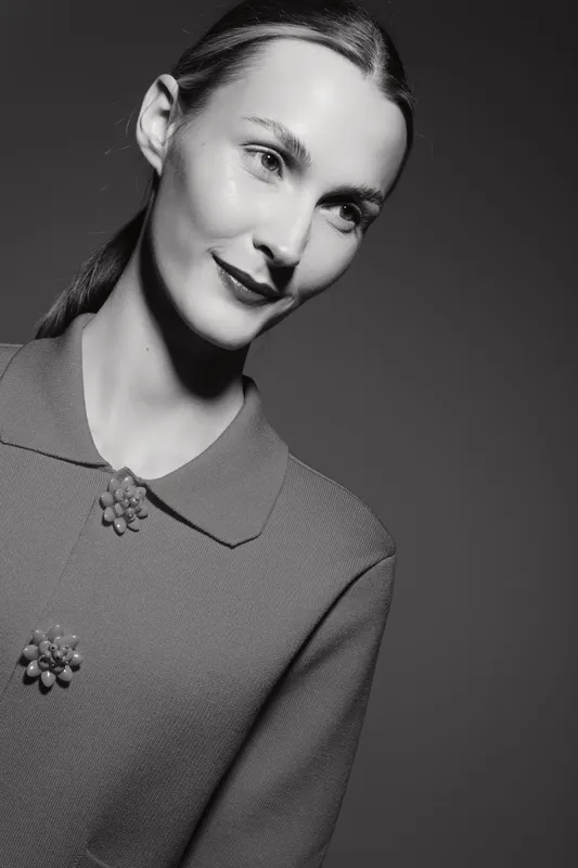 Portrait of a fair skinned caucasian woman with slicked back hair wearing a textured top with decorative buttons. Soft studio lighting highlights her serene expression and smooth skin.