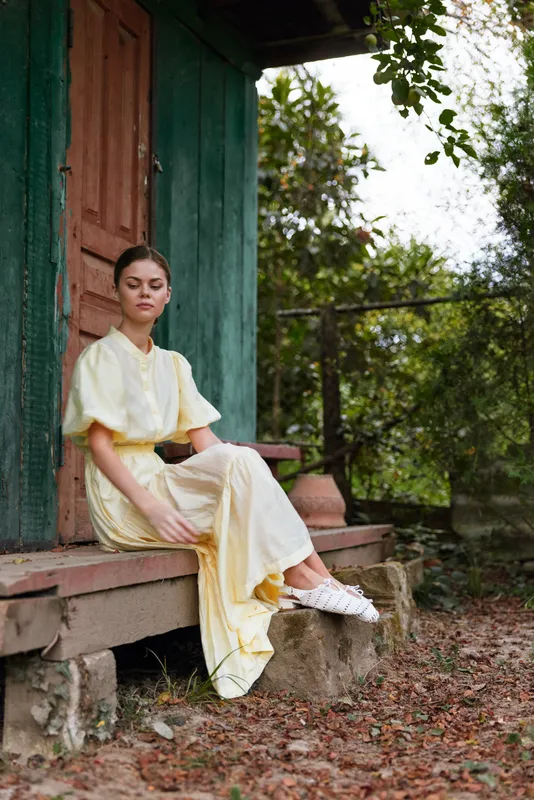 Woman in a yellow dress sitting on a cottage porch in rural nature, vintage portrait showing calm emotion and relaxation with authenticity inclusivity in outdoor scene
