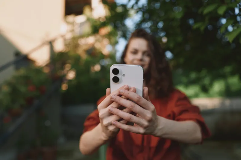 Young woman outdoors holds a smartphone and takes a selfie, smiling softly as she focuses on the screen, with a sunlit garden in the blurred background