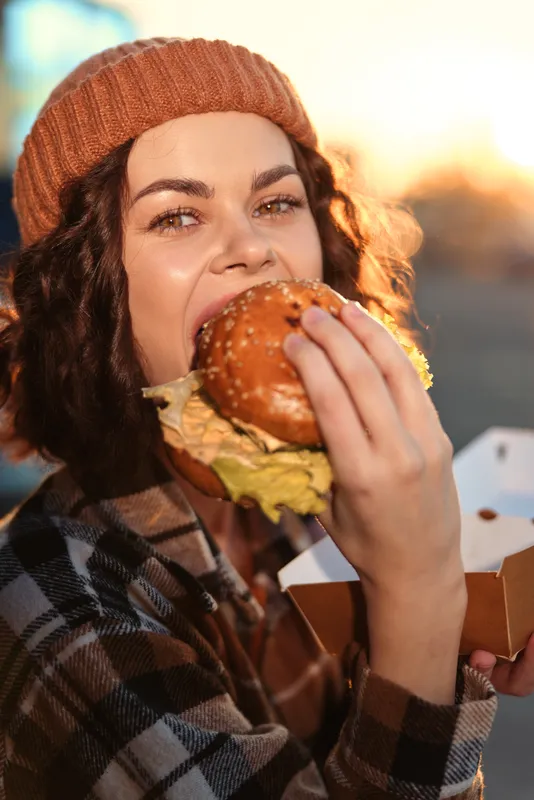 Woman eating burger with smile in autumn beanie, candid takeaway closeup on street at golden hour glow. Authenticity and emotional storytelling for mindful living, modern urban moment.
