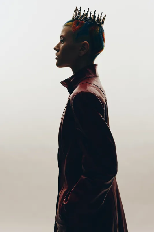 Profile of a woman with colorful braided hair and a spiked crown, wearing a dark leather jacket against a plain light background, showcasing edgy fashion and individuality.