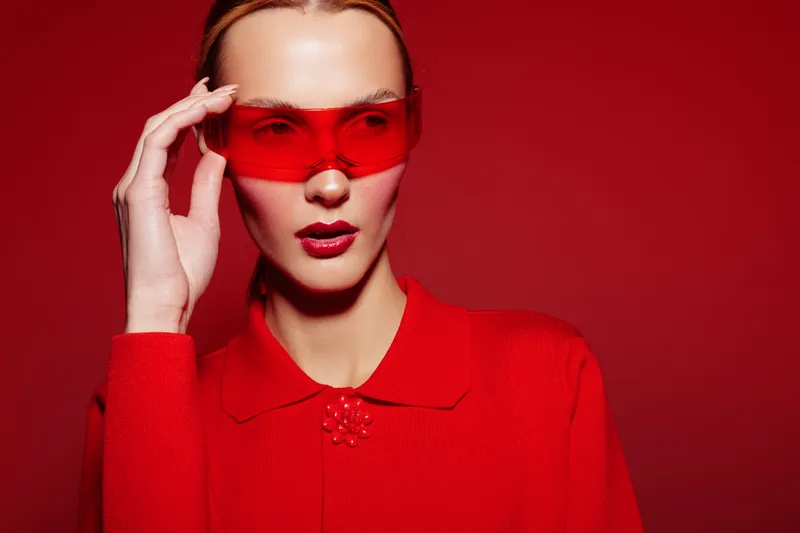 Stylish woman in red sweater and red sunglasses poses against red background. Fashion portrait with bold color contrast and confident expression. Concept of modern style and elegance.