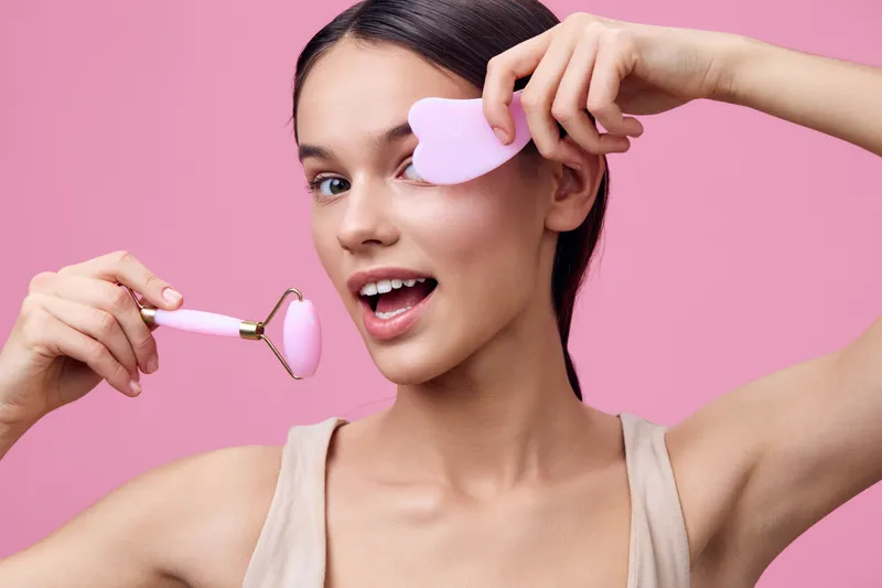 Young woman with a joyful expression using skincare tools against a pink background, promoting beauty and self care routines for glowing skin