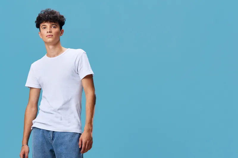 young man wearing casual white t-shirt and blue jeans standing against plain light blue background with a relaxed expression and natural posture