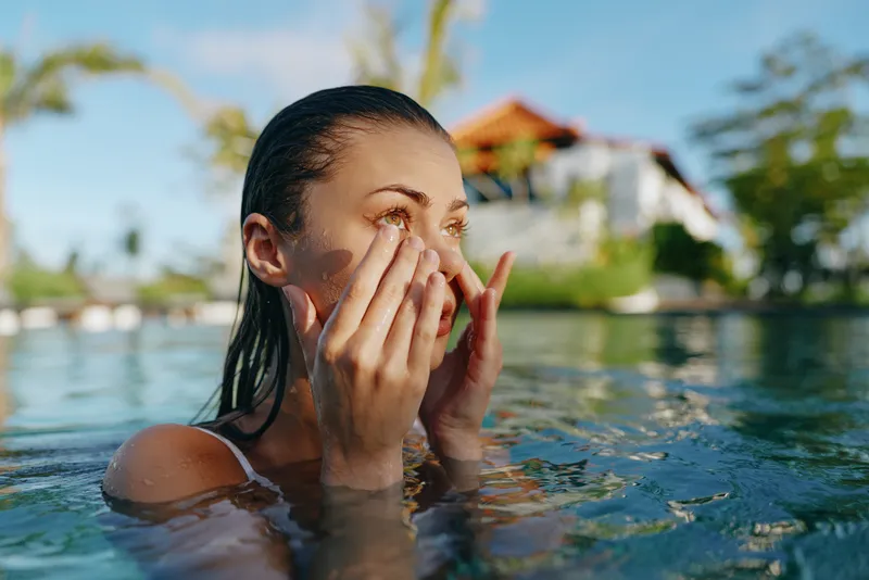 Cheerful woman in a pool, smiling with hands near her face, sunlit water and tropical backdrop, relaxed posture, refreshing summer mood, natural colors, outdoor lifestyle