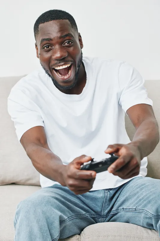 Excited young man playing video games at home, wearing a casual white t shirt and jeans, showcasing joy and enthusiasm in a cozy living room environment