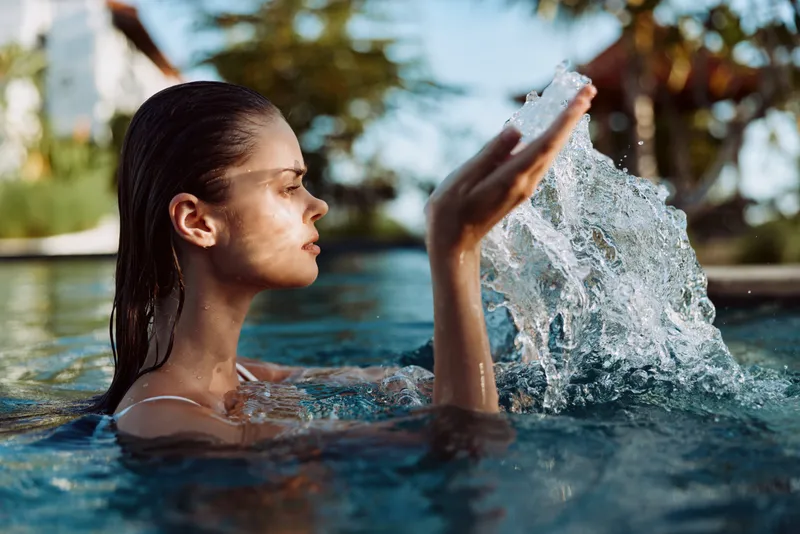 Young woman with wet hair enjoys splashing water in an outdoor swimming pool during a sunny day, expressing relaxation and freshness in a casual swimsuit.