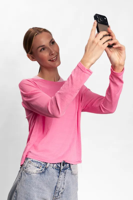 Young woman in pink sweater and jeans takes a selfie with smartphone on white background. Casual style, smiling face, social media concept, technology use and modern lifestyle.
