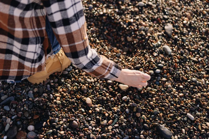 Pebbles and stones dot a sunlit beach as a person kneels, picking small rocks with a cautious touch. Closeup of hand and plaid shirt fabric creates a warm, tactile outdoor scene