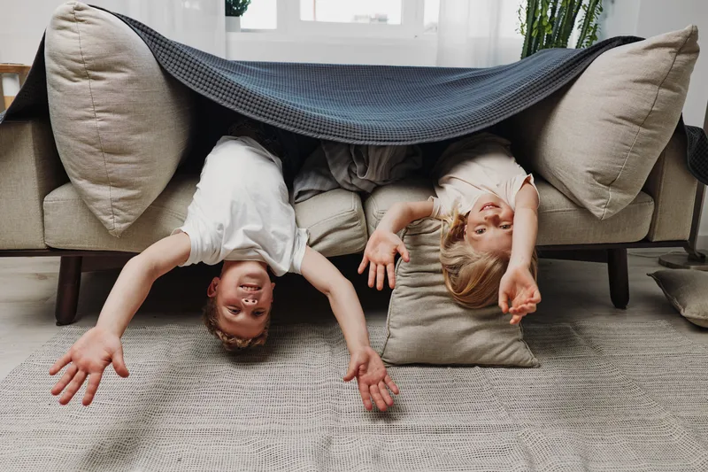 Two happy children lying upside down under a blanket on a couch stretch their arms forward in a cozy living room.
