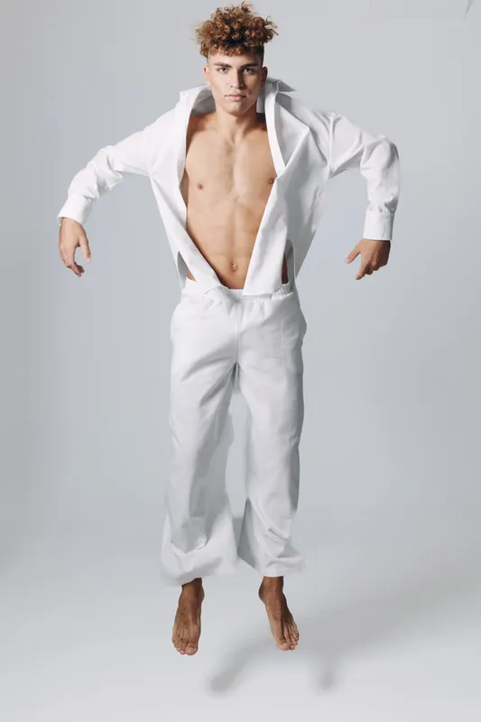 Young man jumping in white loose pants and open shirt with bare chest on gray background. Athletic male model with curly hair in dynamic pose and casual clothing barefoot in studio.