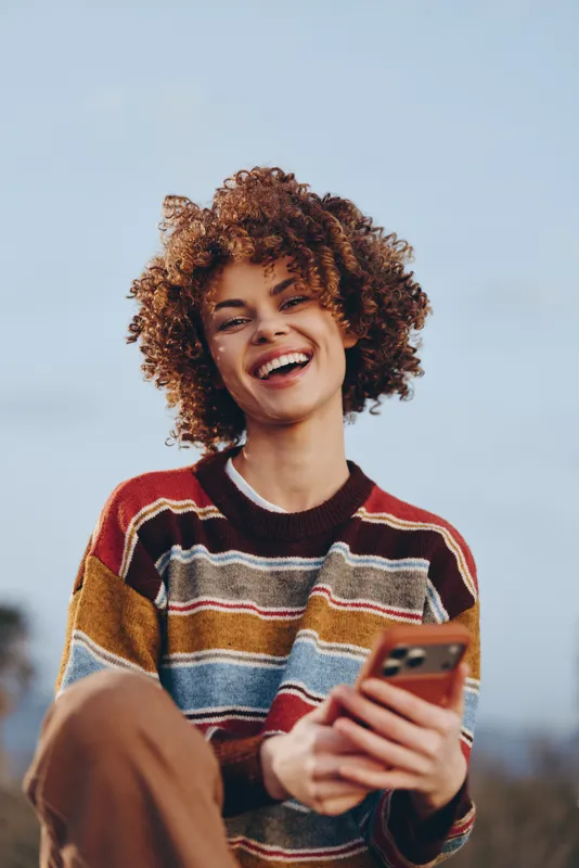 Woman with curly hair smiles outdoors holding smartphone, lifestyle portrait in natural daylight, casual sweater, friendly communication with copy space, soft blurred background open sky