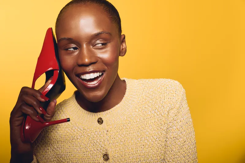 Happy African woman with dark skin smiling and holding a red high heel shoe near her ear. Yellow background and beige sweater create a bright and cheerful concept of fashion and joy.