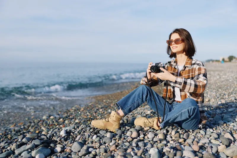 Woman photographer sits on a rocky beach with a handheld camera, enjoying a sunny day by the sea, casual plaid shirt, denim jeans, boots and sunglasses while capturing coastal scenery.
