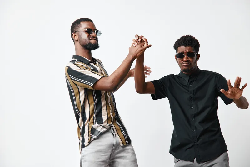 Two young black men with dark skin wearing sunglasses and casual shirts dancing and having fun together on white background in studio.