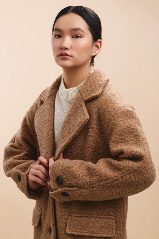 Asian woman wearing a warm brown coat stands against a neutral backdrop, exuding calm confidence and refined style in a minimal portrait.