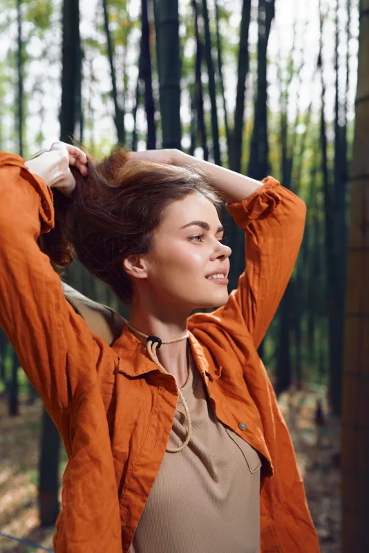 Woman forest portrait, smile and sunlight on her face, casual orange jacket and relaxed pose with hands in hair. Outdoor nature portrait with warm tones and peaceful expression.