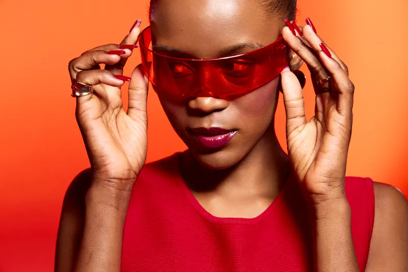 Close-up of a confident dark skinned African woman wearing red sunglasses and red top against orange background. Concept of fashion, style, beauty, and modern accessories for women.